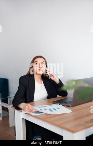 Business Woman Talking on Smartphone While Working at Office Desk With Laptop. Corporate Workplace and Communication Concept. Female Professional. Stock Photo