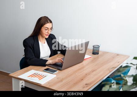 Woman working on laptop at office desk with smartphone and documents. Beautiful businesswoman multitasking in corporate workplace. Female professional Stock Photo