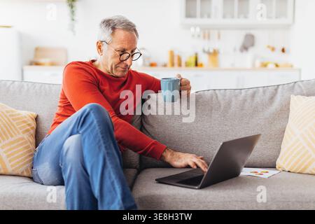 Senior Man Using Laptop at Home While Enjoying a Cup of Coffee in the Living Room Stock Photo