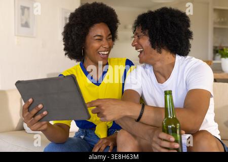 African couple laughing, sharing tablet on beige couch at home, woman in yellow, man holding beer Stock Photo