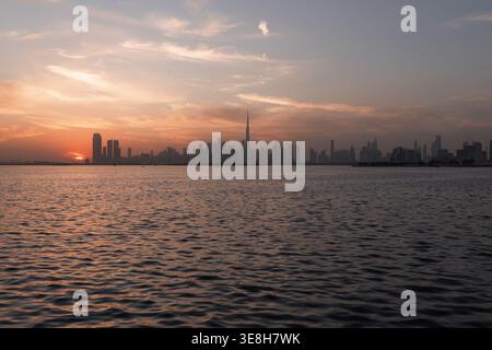 View of the sun setting behind the Dubai skyline, casting a warm glow over the water, with the Burj Khalifa standing tall, Dubai, United Arab Emirates Stock Photo