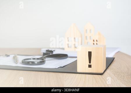 Single exclamation mark on a wooden block. Blurred background with miniature houses, magnifying glass and contract. Real estate warning concept. Stock Photo