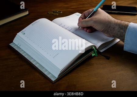 Hand writing appointments in planner on wooden desk with phone and coins, home productivity and time management concept. Stock Photo