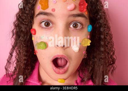 A Surprised Girl with Colorful Candy Stuck on Her Face Against a Pink Background Stock Photo