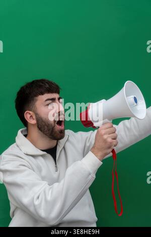 Young man holding and shouting into a megaphone, making an announcement or protesting against a green screen Stock Photo