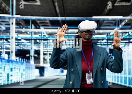 Data center female programmer using VR headset gear to configure server settings. IT specialist in modern high tech server hub uses virtual reality technology to do maintenance tasks on infrastructure Stock Photo