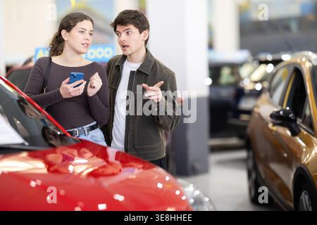 Young couple is standing in a car dealership and looking at the car with gadget in hands Stock Photo