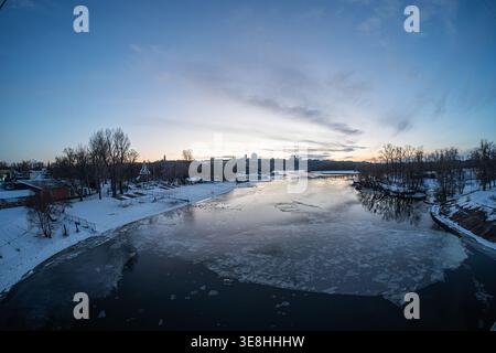 Icy river at dusk with buildings and trees, orange and blue sky with scattered clouds, shimmering reflections on the ice Stock Photo