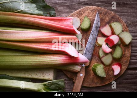 Whole rhubarb stalks with sliced pieces on wooden table, kitchen preparation concept. Fresh organic rhubarb and knife, healthy seasonal ingredient. Top view Stock Photo