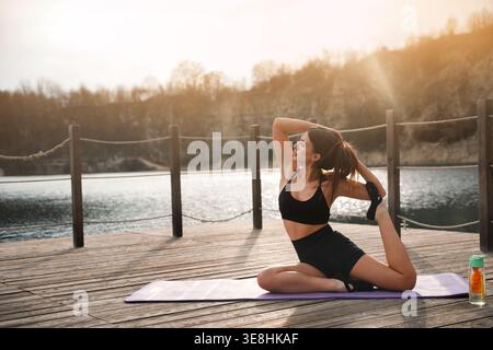 Outdoor yoga. Young woman practicing stretch pose near water. Female improving flexibility and balance. Healthy lifestyle. Stock Photo