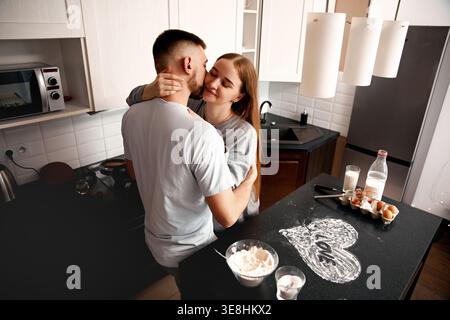 A couple hugs in the kitchen while preparing food. They enjoy their time together as they cook and connect in the warm setting of their home. Stock Photo