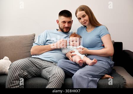 A young couple sits on a couch holding their baby. They are relaxed and smiling. The setting is a living room with soft light. The family shares a spe Stock Photo