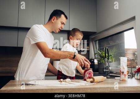 Father and son work together in the kitchen. The father helps the son spread jam on dough while they prepare snacks. The kitchen is bright and organiz Stock Photo