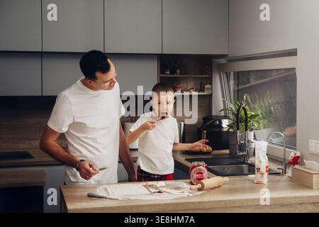 Father and son are working in a modern kitchen. The son is tasting food while the father watches. They are surrounded by kitchen tools and ingredients Stock Photo