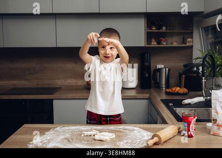 Child stands in kitchen and rolls dough on counter, showing excitement while preparing food. Various kitchen tools and ingredients are present in a br Stock Photo
