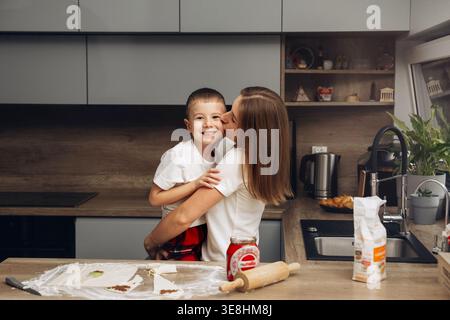 A mother holds her son in the kitchen as they prepare to bake cookies. The kitchen has cooking items on the counter and a friendly atmosphere during f Stock Photo