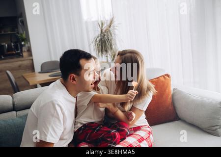 Two parents and a child spend time together on a couch while the child playfully holds a lollipop. They share smiles and laughter in a cozy living roo Stock Photo