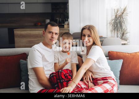 Family enjoys a cozy time together on the couch in their living room. The child is happily holding a lollipop while wearing matching pajamas with the Stock Photo