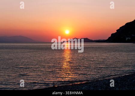 Sunset over calm sea with mountain silhouettes and golden reflection on water in Greece Stock Photo