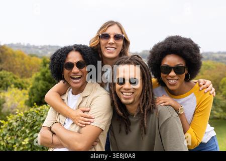 Diverse friends posing on park hill, wearing sunglasses, wristwatch, bracelet, hugging Stock Photo