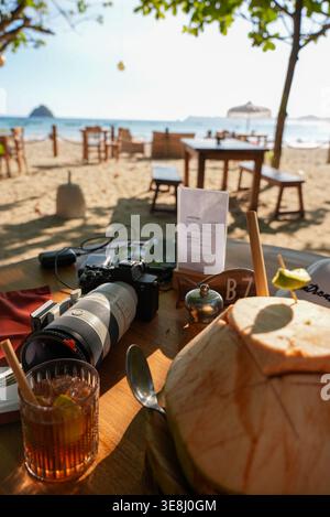 Camera, glass with drink and coconut on table with beach chairs and ocean visible in the background. Stock Photo