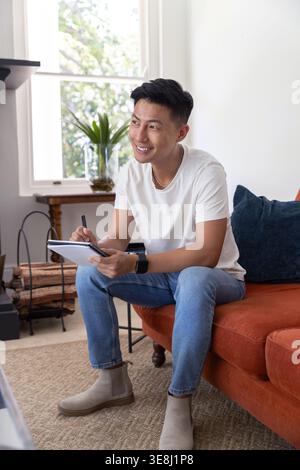 Asian man sitting on orange sofa holding spiral notebook and pen in living room wearing smartwatch Stock Photo