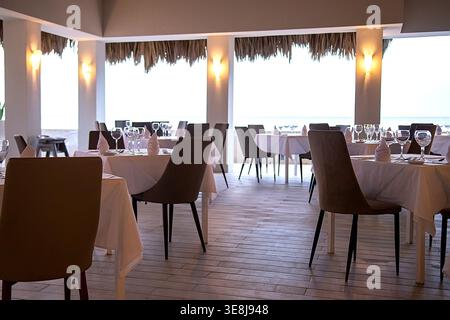 Elegant interior of a seafood restaurant with high thatched ceiling, large artistic fish-shaped chandelier, wooden beams, set white tables with linens Stock Photo