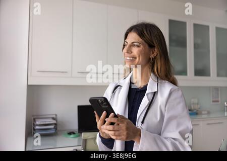 Smiling Latina woman therapist in white coat holding smartphone Stock Photo