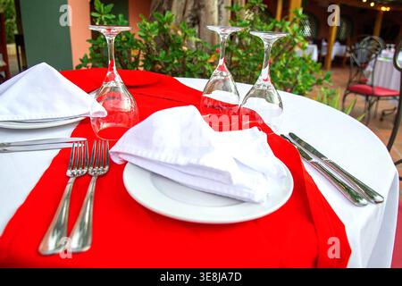 Elegant table setting with red runner, white plates, folded napkins, cutlery and wine glasses on round table amid garden patio. Captures authentic ser Stock Photo