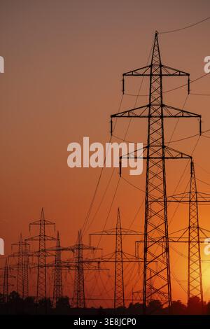 Silhouette of several high-voltage power lines and electricity pylons against a glowing orange and red sunset sky in Germany Stock Photo