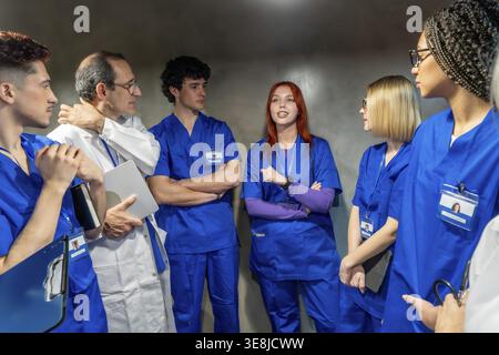 Medical professionals including doctors and nurses, wearing blue scrubs and a lab coat, standing and actively engaged in a serious discussion about he Stock Photo