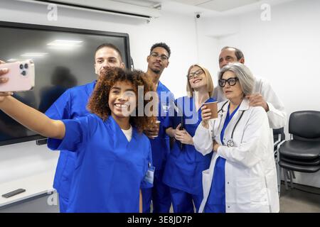 Group of smiling multi ethnic doctors, nurses, and medical personnel posing for a selfie using a cell phone, symbolizing joyful collaboration, modern Stock Photo