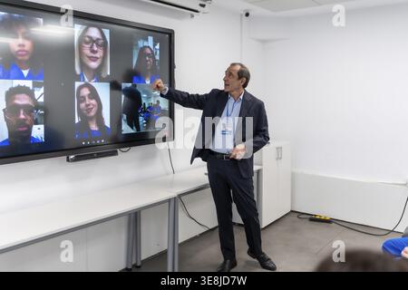 Teacher leads virtual class, pointing to large interactive screen displaying a grid of remote students in a video conference, demonstrating modern dig Stock Photo