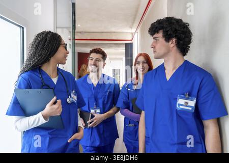 Diverse group of young medical students and healthcare professionals in blue scrub uniforms walking and talking in a modern hospital or clinic hallway Stock Photo