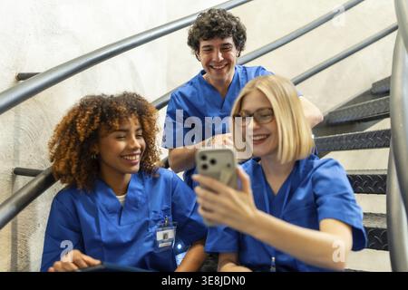Group of diverse medical students or healthcare professionals wearing blue scrubs, smiling and interacting while looking at a mobile phone on a stairc Stock Photo