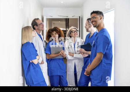 Happy multiethnic medical team of doctors and nurses in scrubs and lab coats standing in a clinic corridor, smiling and exchanging ideas while using a Stock Photo