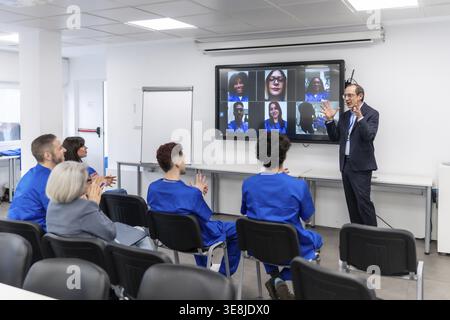 Experienced professor leads hybrid medical seminar, engaging in person students while remote participants appear on a large monitor for interactive he Stock Photo