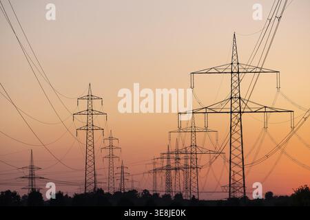 Silhouette of several high-voltage power lines and electricity pylons against a glowing orange and red sunset sky in Germany Stock Photo