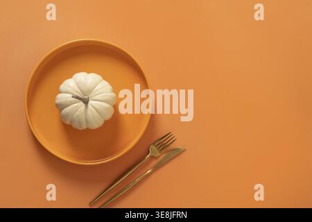 Autumn and Thanksgiving Day table setting with white small pumpkin, orange empty plate with golden cutlery on orange background. Concept catering. Top Stock Photo