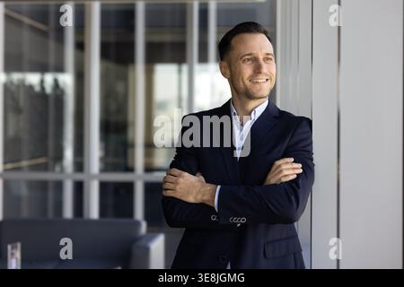 Confident businessman stands with folded arms, looks into distance thoughtfully Stock Photo