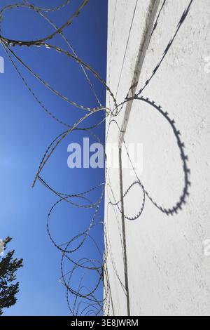 Barbed wire on white wall and blue sky with shadows from bright sun. Stock Photo