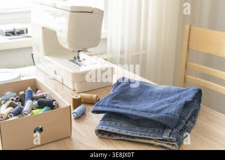 Sewing machine on wooden table beside folded blue jeans and an open box of colorful thread spools, sunlight from window illuminating workspace Stock Photo