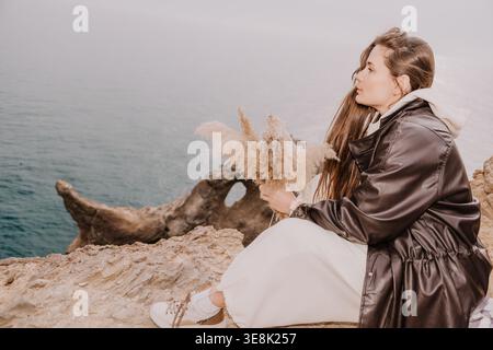 Woman cliff sea sitting on a rocky coast holding dried pampas grass while looking at the misty morning ocean landscape Stock Photo