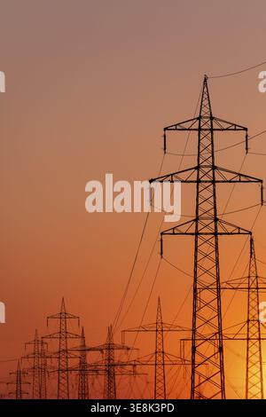 Silhouette of several high-voltage power lines and electricity pylons against a glowing orange and red sunset sky in Germany Stock Photo