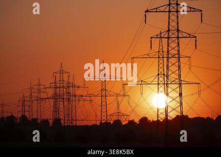 Silhouette of several high-voltage power lines and electricity pylons against a glowing orange and red sunset sky in Germany Stock Photo
