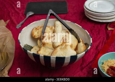 A large striped ceramic bowl filled with assorted small flatbreads and breadsticks sits on a deep red velvet tablecloth, with metal serving tongs rest Stock Photo
