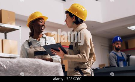 Multiethnic team of women checking stock of goods in warehouse, planning industrial distribution with packages. Two people in overalls and hardhat using digital tablets. Handheld shot. Stock Photo