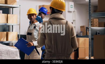 Multiethnic group of people in overalls checking goods stacked in boxes on storage room shelves, using inventory list. Depot workers planning order distribution and shipment, industrial cargo. Stock Photo