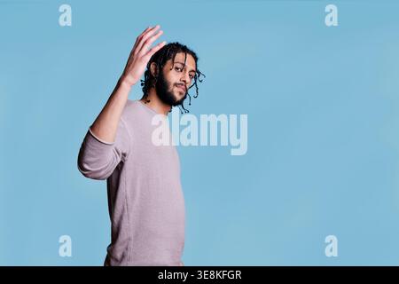 Handsome arab man raising hand and looking at camera with confident facial expression. Smiling young adult person with black braided hairstyle portrait on blue background Stock Photo