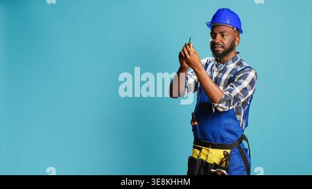 Carpenter in overalls holding screwdriver in studio, using handy instrument to screw nails. Construction engineer working with manual utility tool, screwing something over blue background. Stock Photo